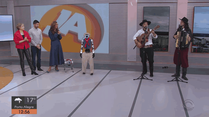 GIF em plano aberto do Jornal do Almoço: o robô Tenório e três apresentadores dançam juntos no estúdio, ao lado de dois músicos e de um cão-robô.